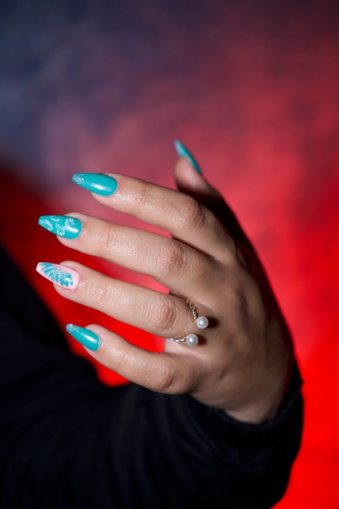 Close-up of a hand with turquoise nail art, adorned with rings and pearls, on a vibrant background.