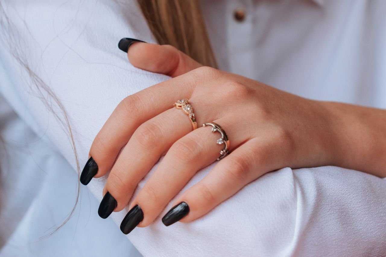 Close-up of hands with black nail polish and elegant rings against a white shirt backdrop.